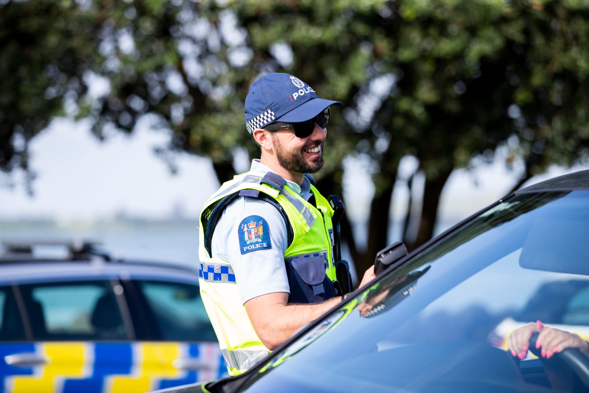 A smiling New Zealand Police officer in a high-visibility vest talking to a driver beside a patrol car