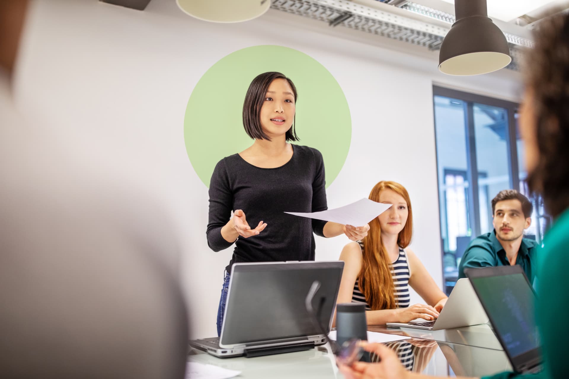 A woman standing and presenting to colleagues gathered around laptops in a bright office