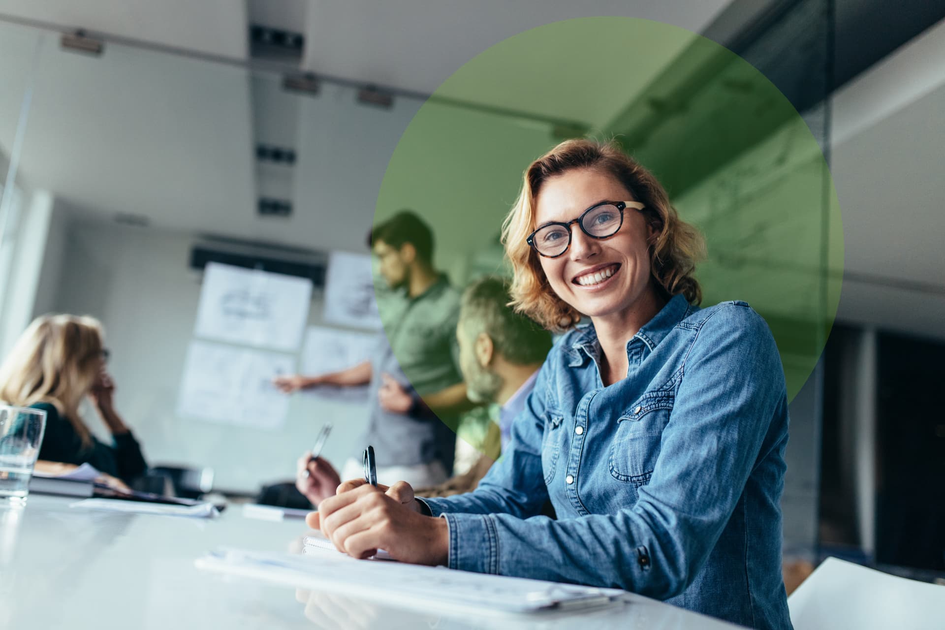 A smiling woman with glasses taking notes at a bright meeting room table while colleagues talk in the background