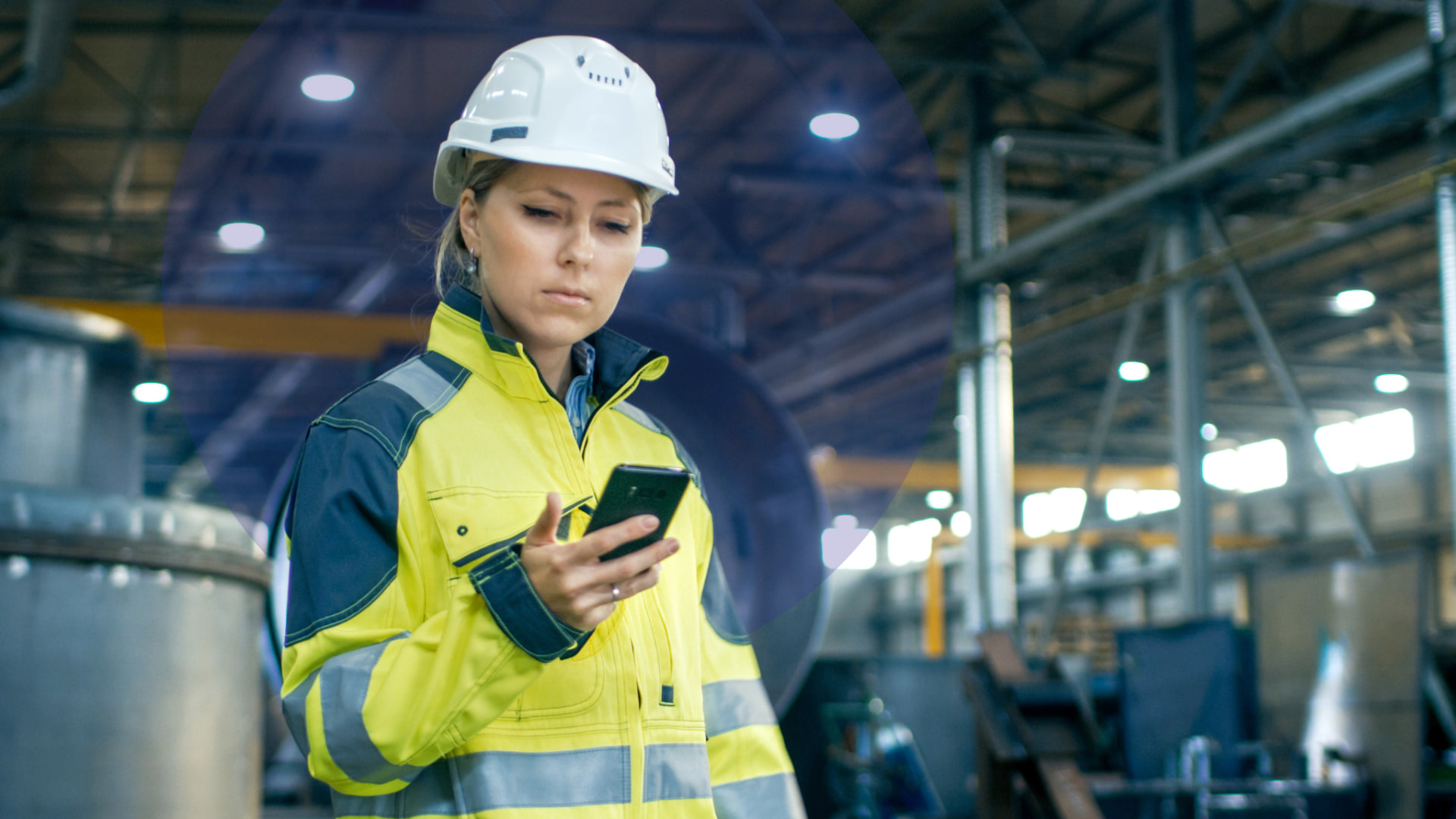 A woman in a high-visibility jacket and hard hat checking her phone in an industrial workplace