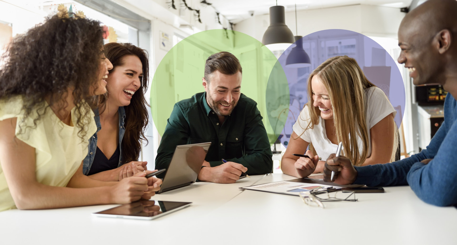 Five colleagues laughing together around a table in a bright office breakout space
